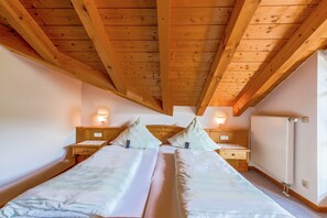 A bedroom with two beds, wooden beams, nightstands, wall-mounted lights, and a radiator.