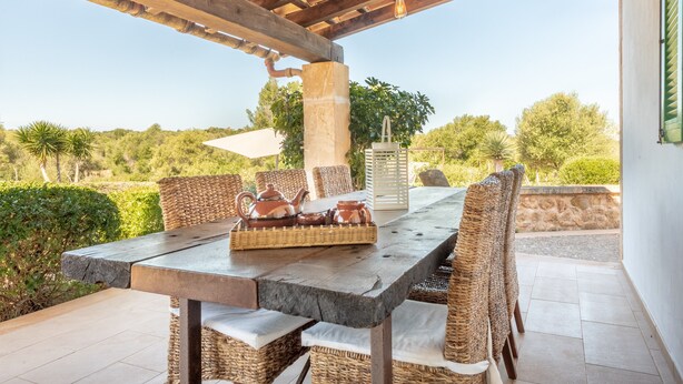 Een terras buiten met een eettafel, rieten stoelen, een stenen pilaar en uitzicht op het omliggende landschap.