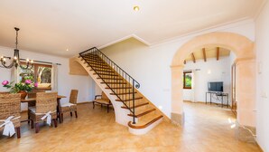 A spacious living area with a wooden staircase, a dining table with chairs, and a TV in the background. - Holiday Home "Cas Batlet Antonia" with Pool, Terrace, Sea & Mountain View (S'HORTA)