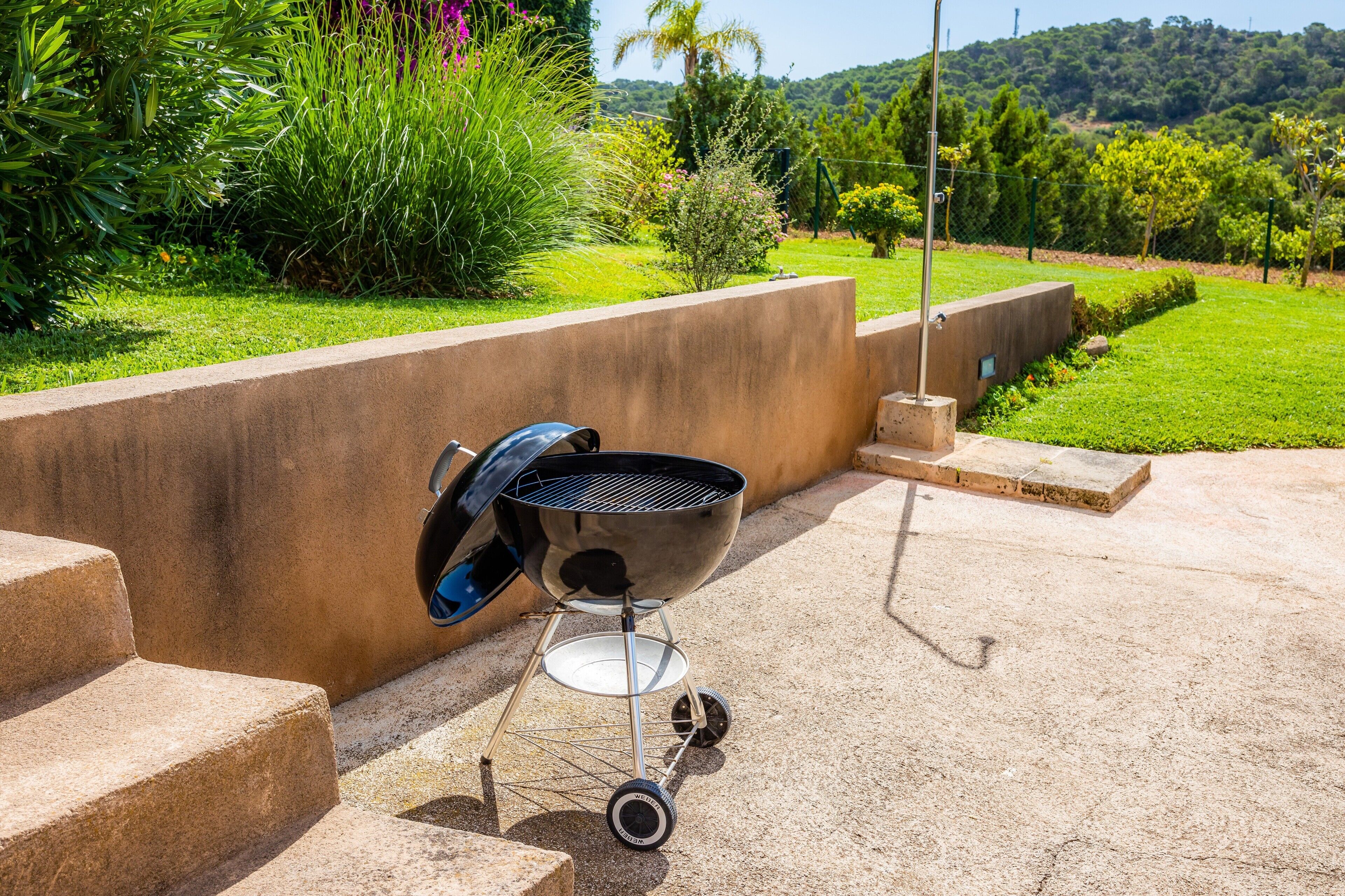 Un barbecue installé sur une terrasse en béton, accessible par des marches, entouré de verdure et offrant une vue sur des collines.