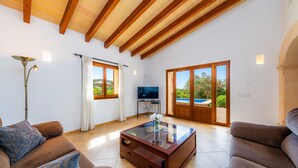 A spacious living room with a high ceiling, wooden beams, a glass coffee table, and a television. - Holiday Home "Cas Batlet Antonia" with Pool, Terrace, Sea & Mountain View (S'HORTA)