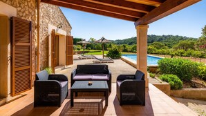 A covered patio with wicker furniture, a pool, and a view of a lush landscape. - Holiday Home "Cas Batlet Antonia" with Pool, Terrace, Sea & Mountain View (S'HORTA)