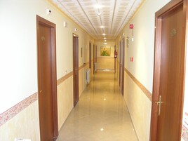 Un pasillo largo y estrecho con puertas de madera a ambos lados, suelo de baldosas e iluminación empotrada en el techo.