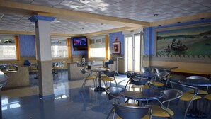 A spacious indoor area with multiple tables and chairs, a television mounted on the wall, and a mural depicting a coastal scene.