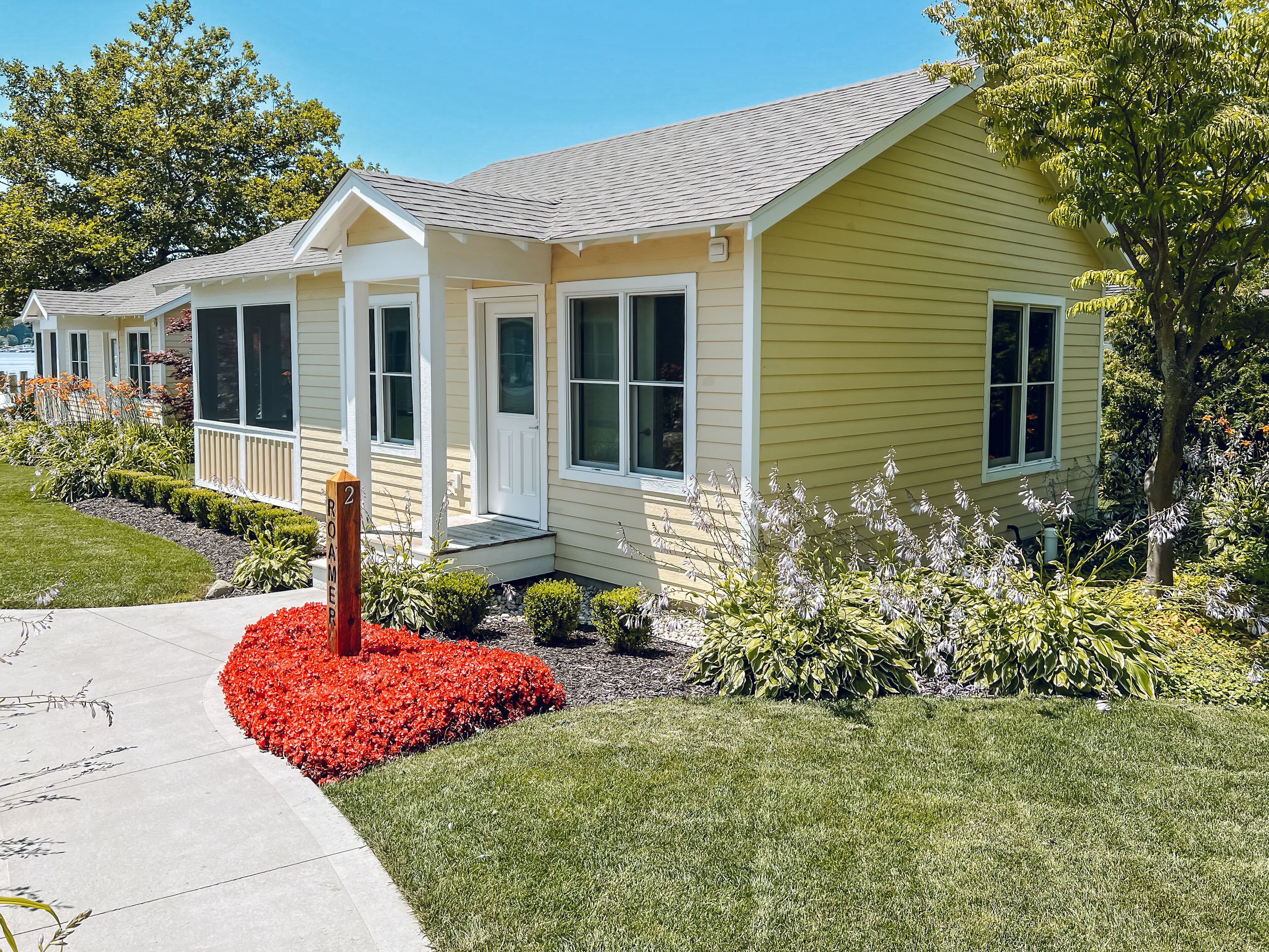 A single-story house with a front porch, a small garden with red flowers, and a mailbox labeled "2 ROAMER".