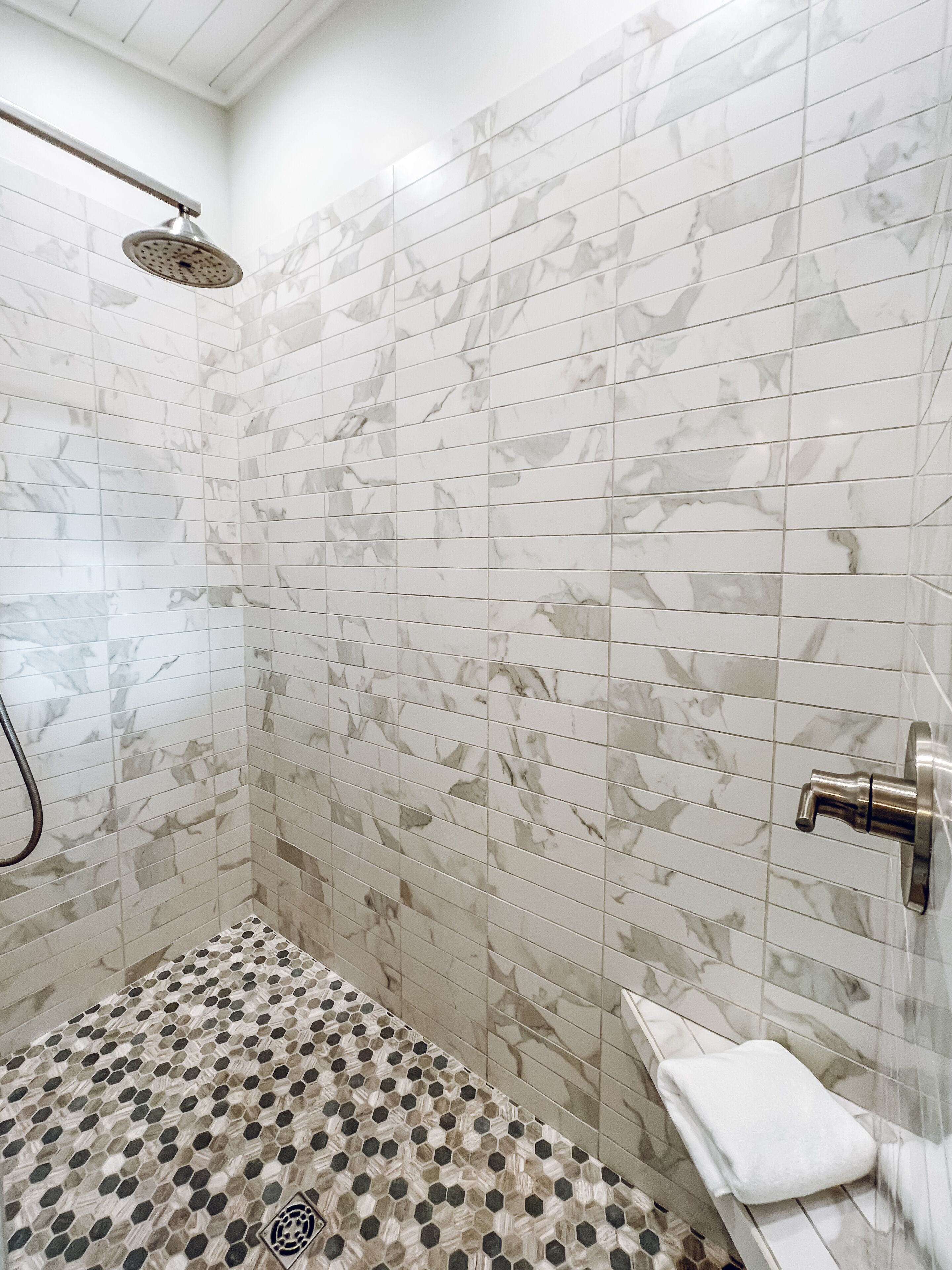 A shower with a rain showerhead and a handheld showerhead, a tiled floor with a hexagonal pattern, and a white towel on a shelf.