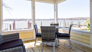 A covered outdoor seating area with wicker chairs and a table, overlooking a body of water with docks and a distant shoreline. - Old Pike Cottages (Saugatuck)