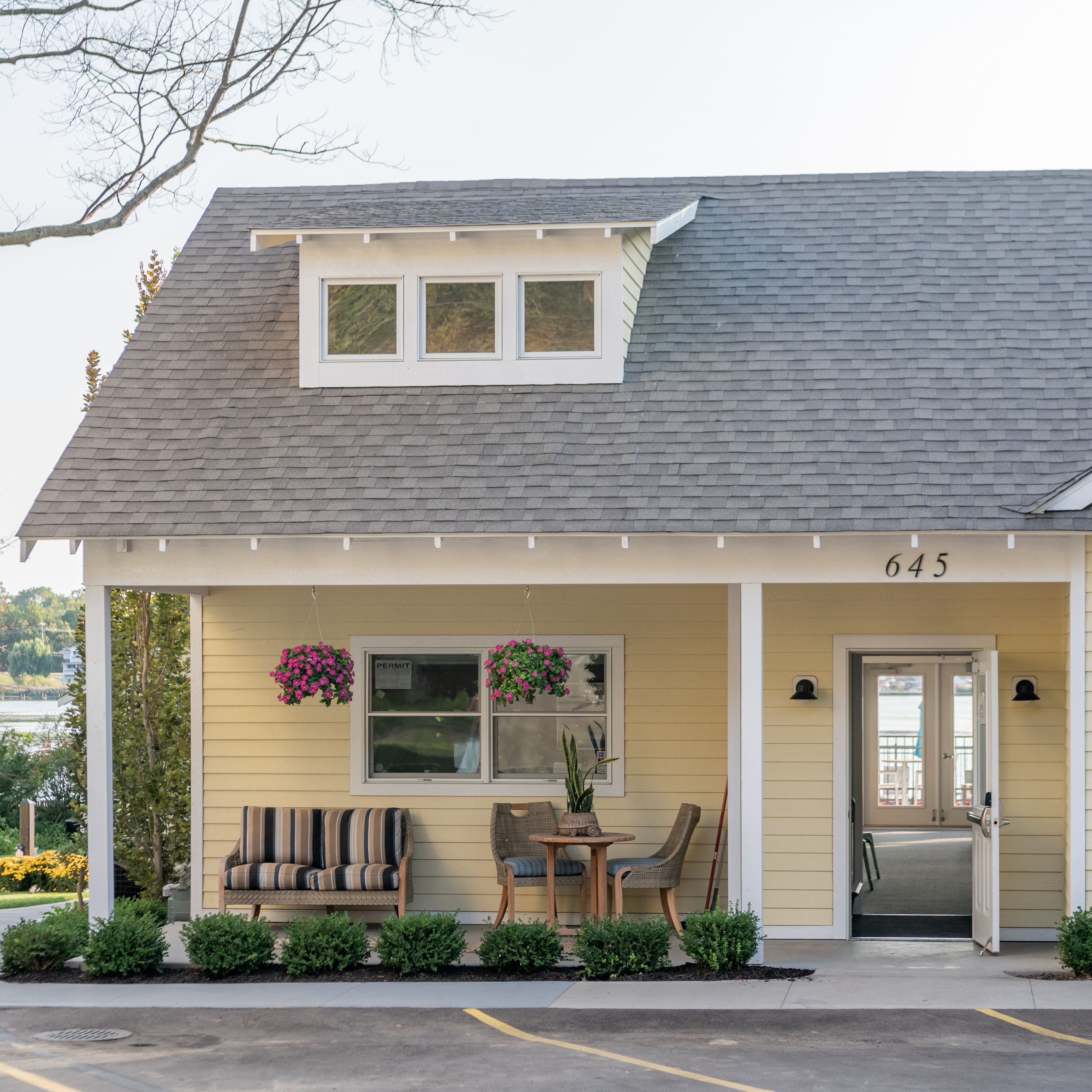 A single-story house with a gabled roof, a front porch with a bench and chairs, a small table, and a door.