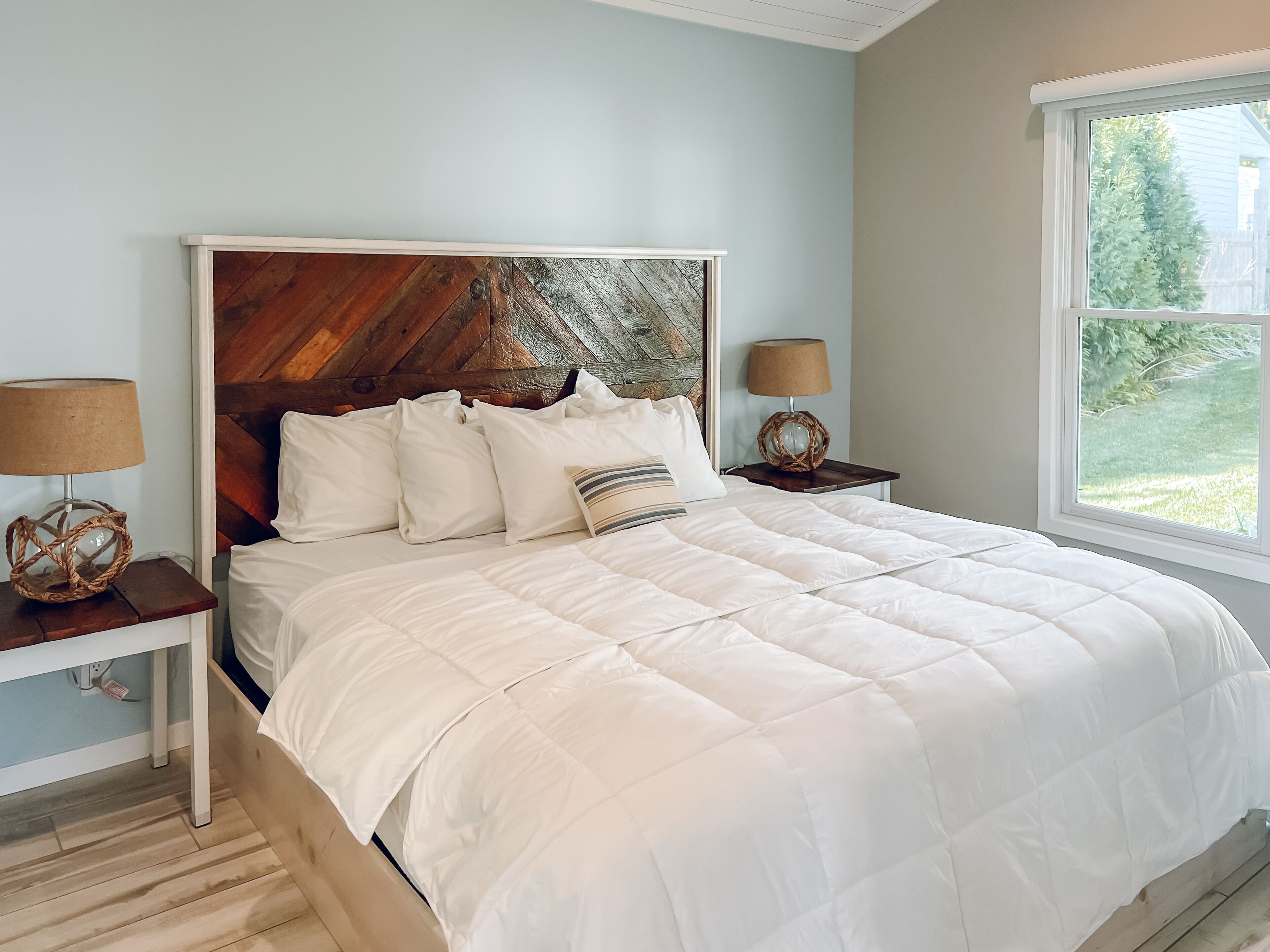 Une chambre à coucher avec une tête de lit en bois, une literie blanche, deux tables de chevet avec des lampes et une fenêtre donnant sur un paysage verdoyant.