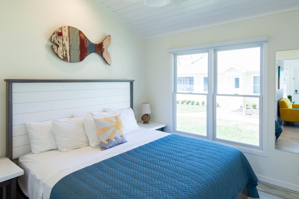A bedroom with a bed, white bedding, a blue patterned bedspread, a wooden fish-shaped wall decoration, a nightstand with a lamp, and a large window with a view of the outside.