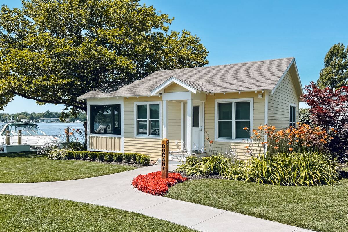 A single-story house with a front porch, a small garden with red flowers, and a dock with boats in the background.