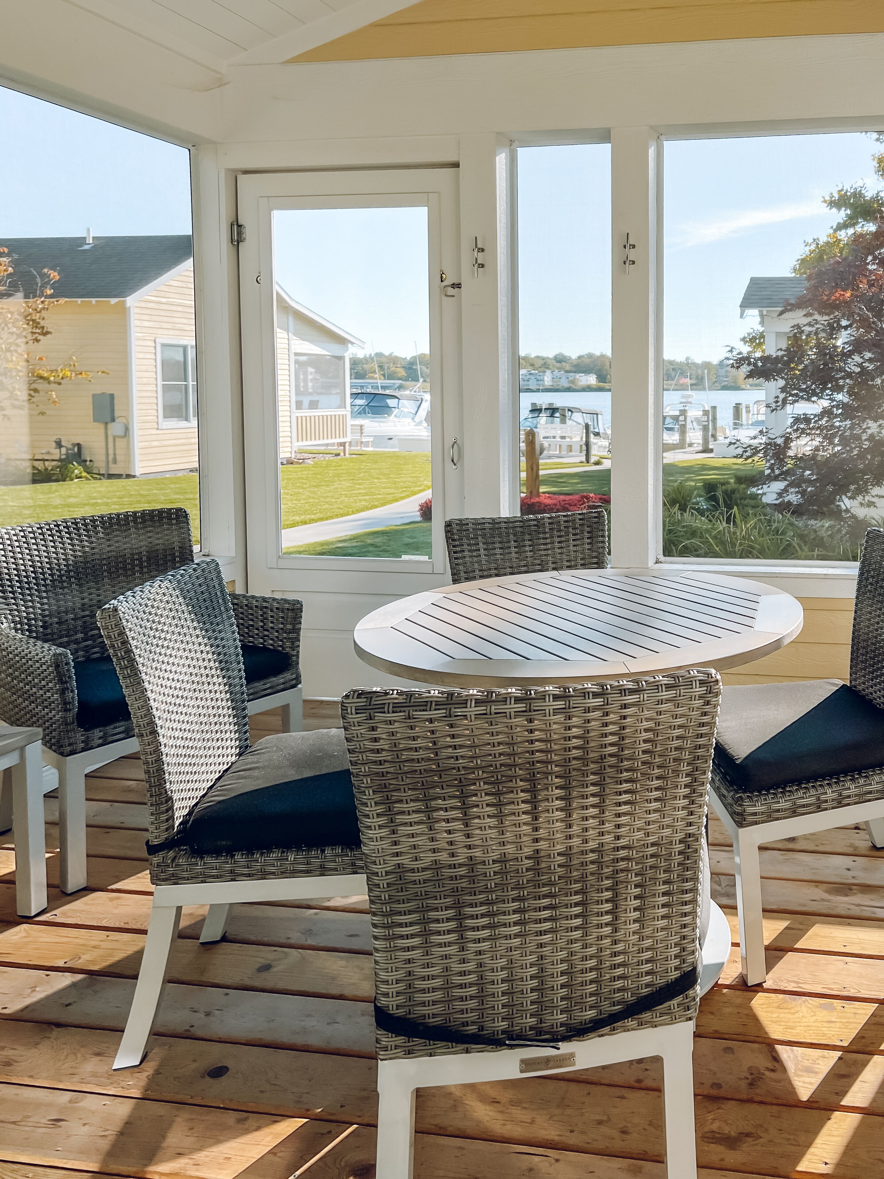 A covered outdoor seating area with wicker chairs and a round table, overlooking a marina with boats and houses.