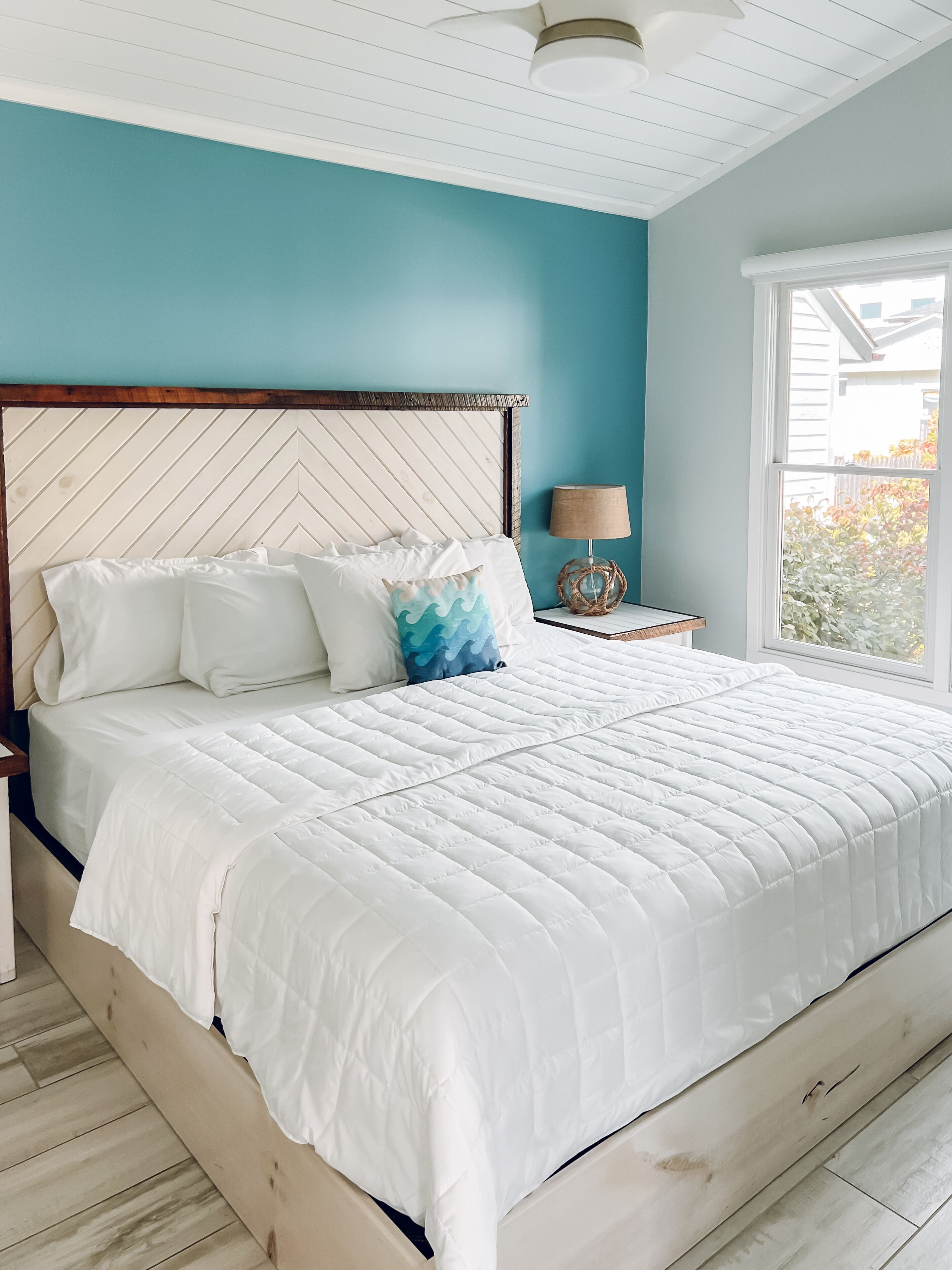 A bedroom with a large bed, a wooden headboard, a nightstand with a lamp, and a window with a view of greenery.