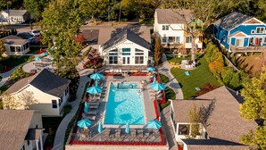 An aerial view of a residential area with a large swimming pool surrounded by lounge chairs and umbrellas, a well-maintained lawn, and houses with different architectural styles. - Old Pike Cottages (Saugatuck)