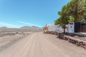 A desert road leading to a building with a stone wall and a tree.