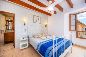 A bedroom with a bed, bedside lamps, a wooden ceiling, a window, and a bathroom with a mirror and sink. - Townhouse with a view — Casa Eliasneil (Andratx)