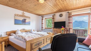A wooden ceiling, a bed with floral bedding, a TV, a chair, a sofa, a window with curtains, and a mountain view. - Vacation on a Farm - Fantastic Holiday App with Balcony, Garden, Lake & Mountain View (Schliersee)