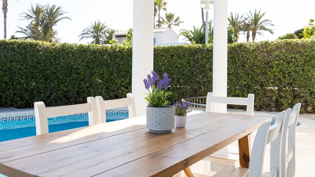 A wooden table with two potted plants, white chairs, and a pool in the background. - Air-Conditioned Holiday Home on the Beach with Pool, Terraces & Wi-Fi (Ciutadella de Menorca)