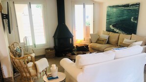 Una sala de estar con un sofá blanco, una silla de mimbre, una mesita redonda, una chimenea negra y un cuadro grande en la pared.
