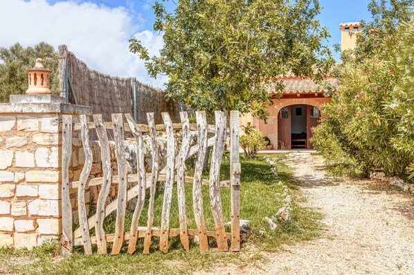 Un muro de piedra rústico, un portón de madera, un sendero que lleva a un edificio con entrada arqueada y una zona arbolada.