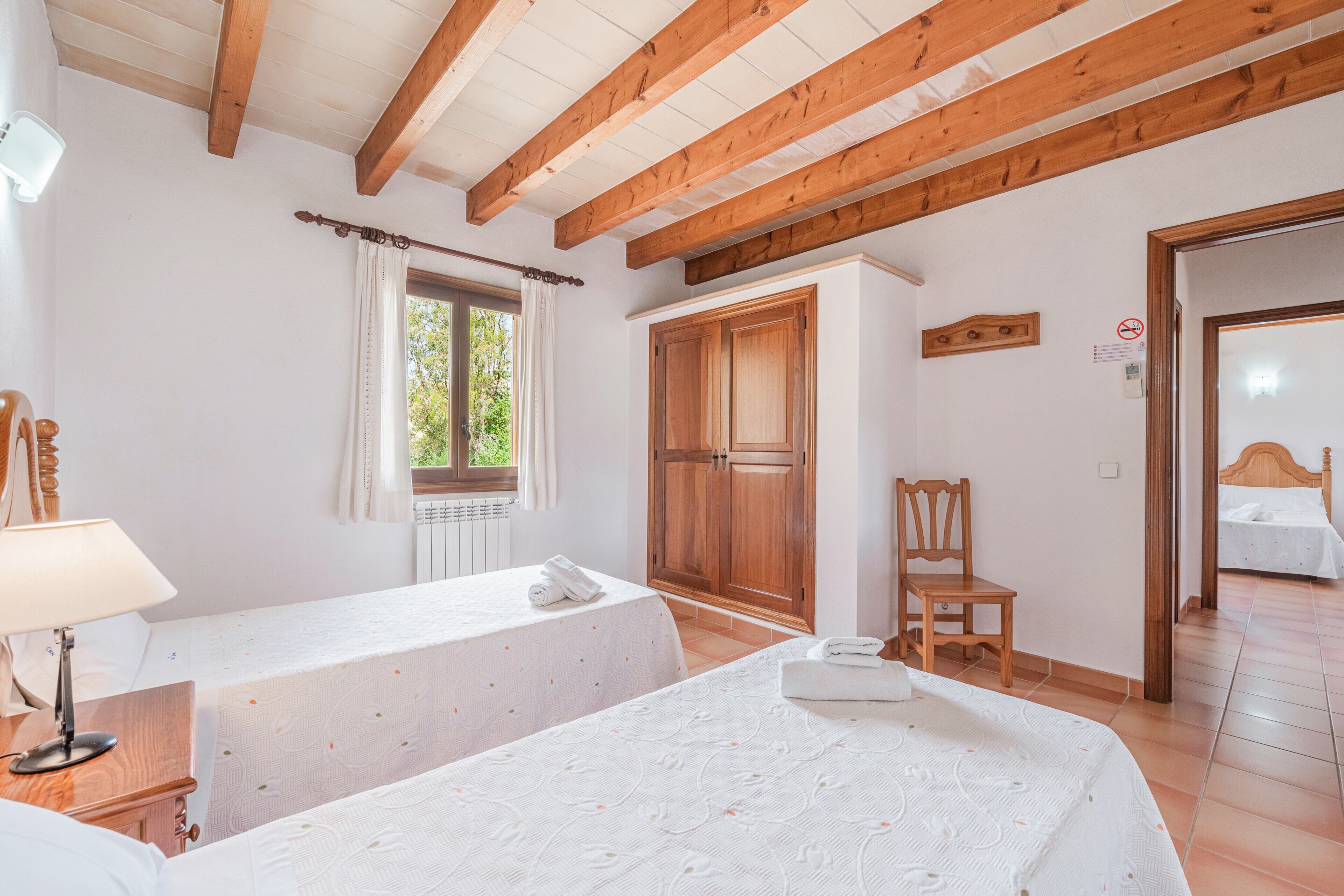 A bedroom with two beds, wooden beams, a window with curtains, a wooden wardrobe, and a chair.