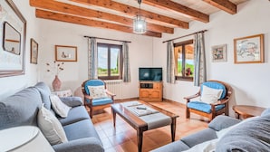 A living room with a sofa, armchairs, a coffee table, a TV, wooden beams, and framed pictures on the walls. - Peaceful location with pool - Villa Can Jaume (Pollença)
