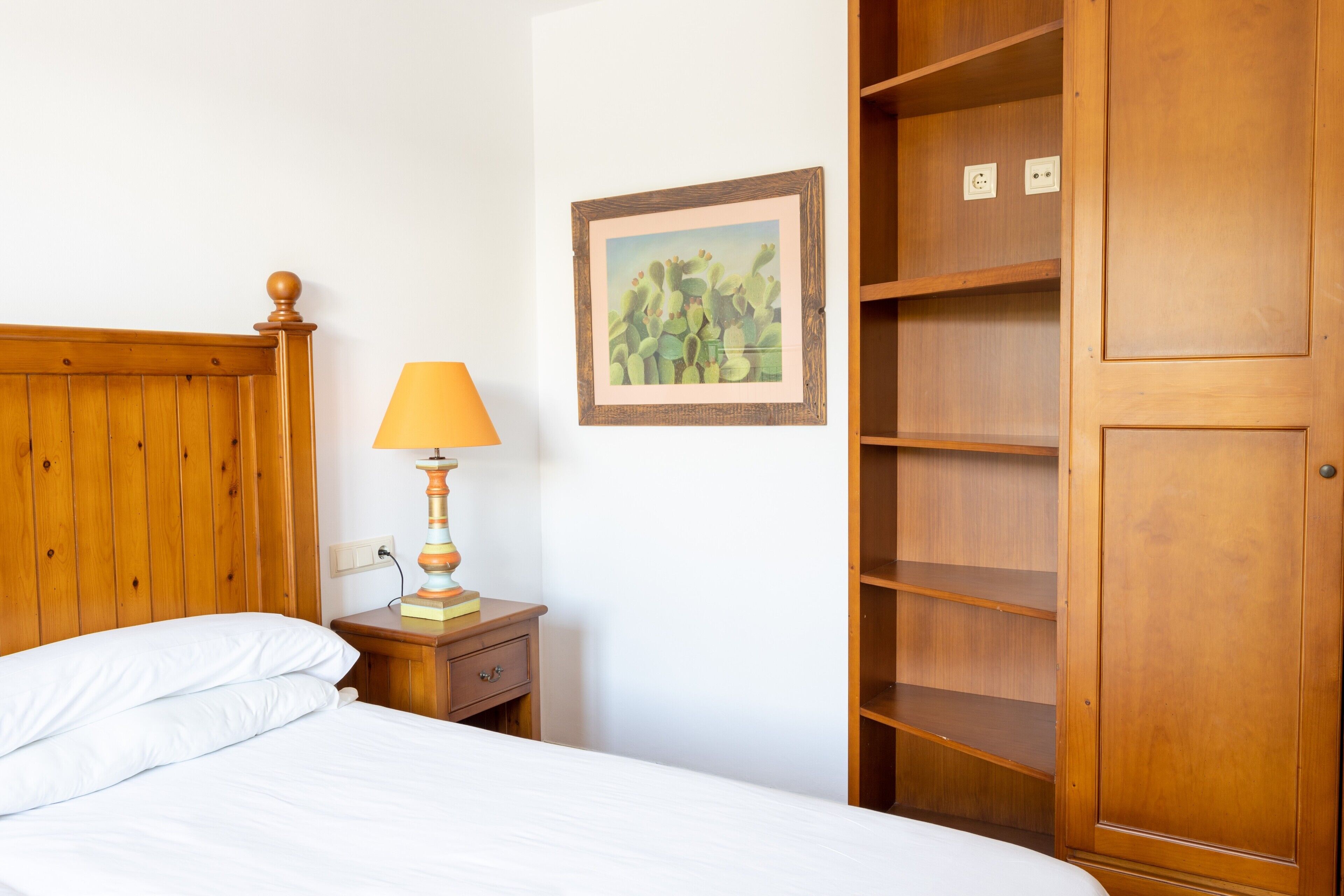 A bed with white linens, a wooden headboard, a nightstand with a lamp, a framed picture on the wall, and a wooden wardrobe.