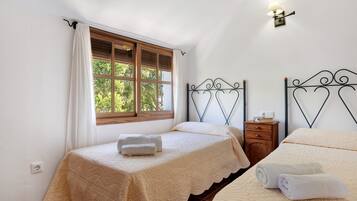 A bedroom with two beds, a window with curtains, a nightstand, and a wall-mounted lamp.