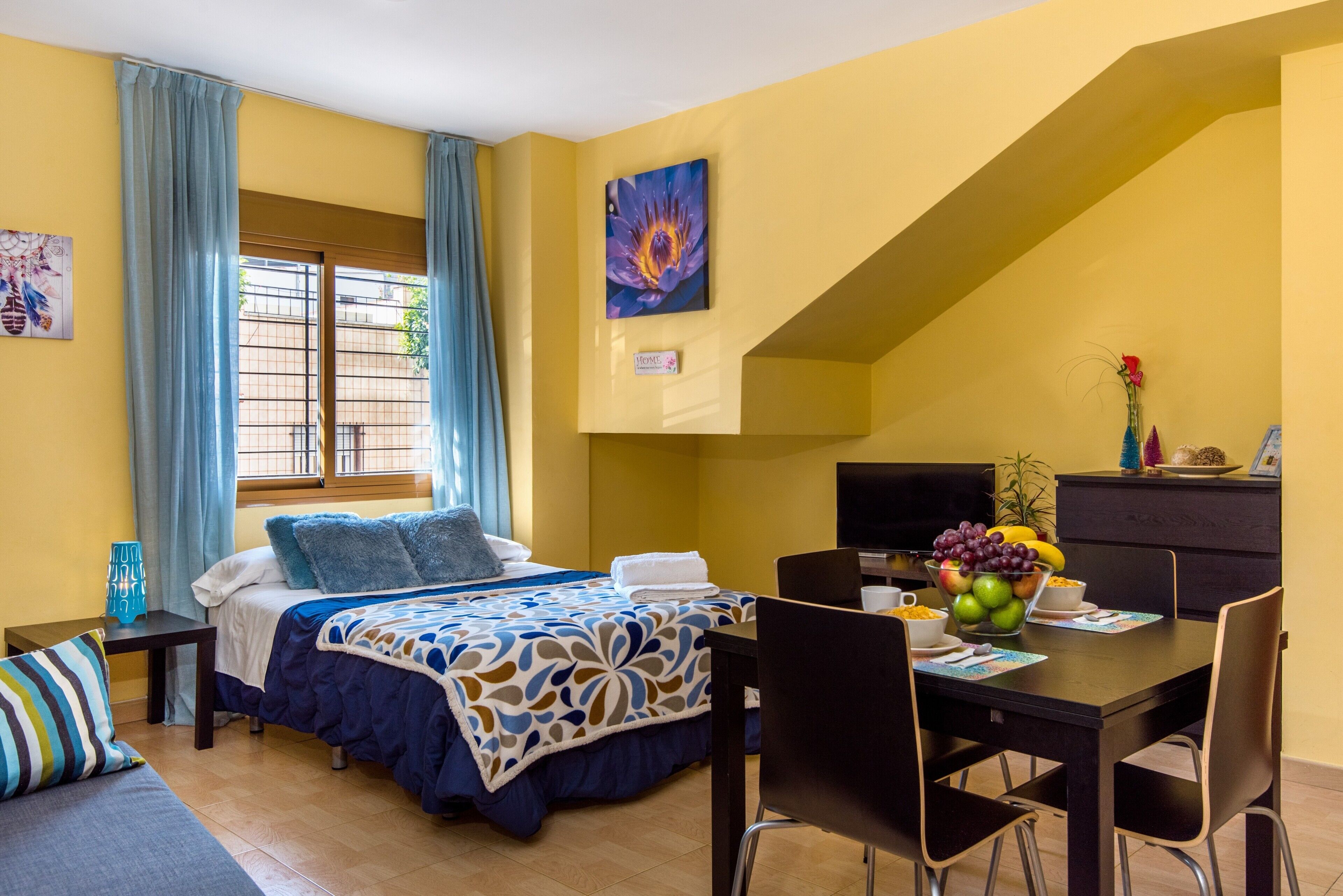 A hotel room with a bed, a desk, a chair, a television, and a dining table with a fruit bowl.