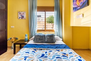 A room with a bed, a window with curtains, a vase with flowers, and a painting on the wall.