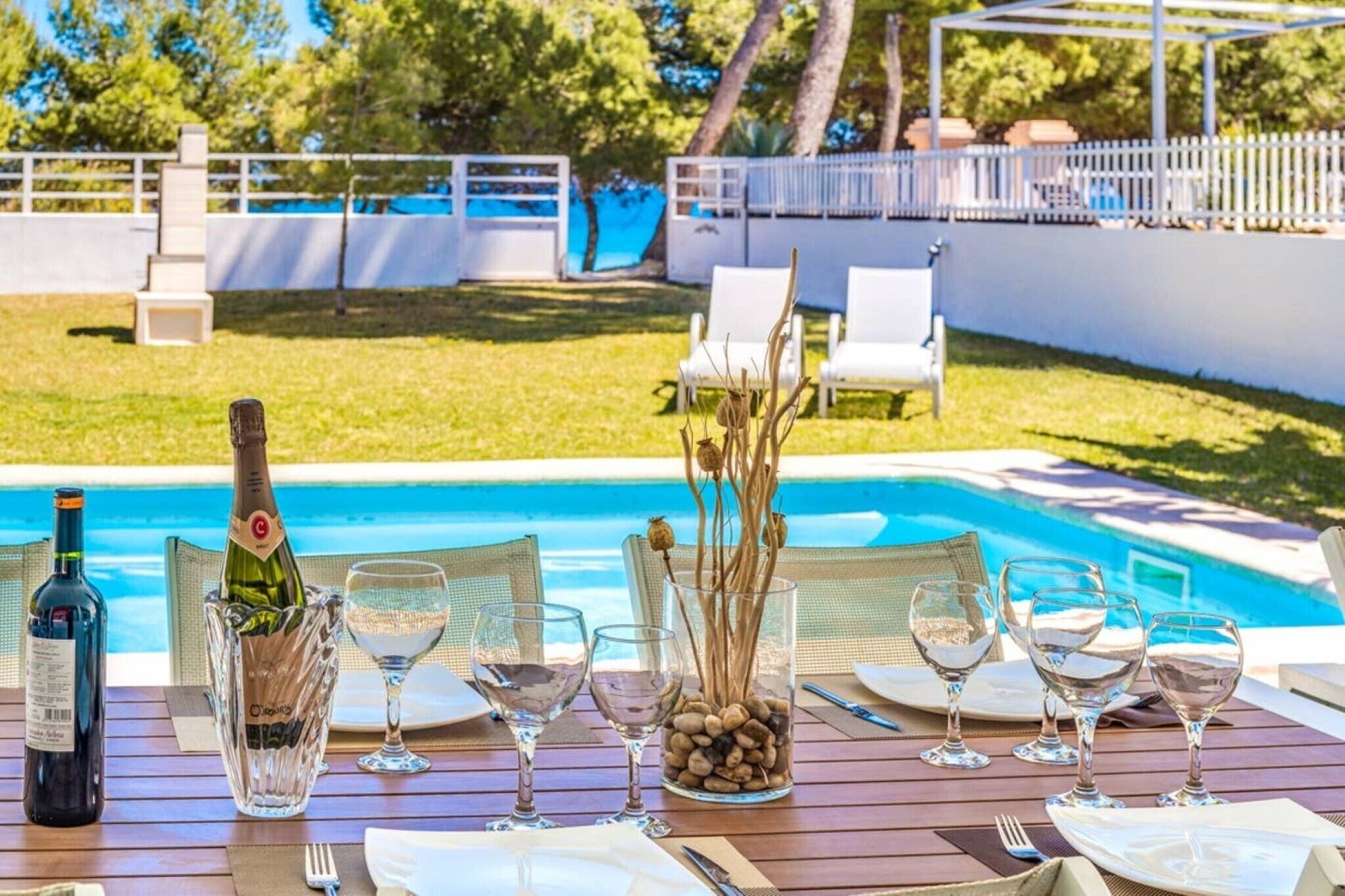 Un área de comedor al aire libre con vista a la piscina, sillas blancas y una mesa para dos con copas de vino y una botella.