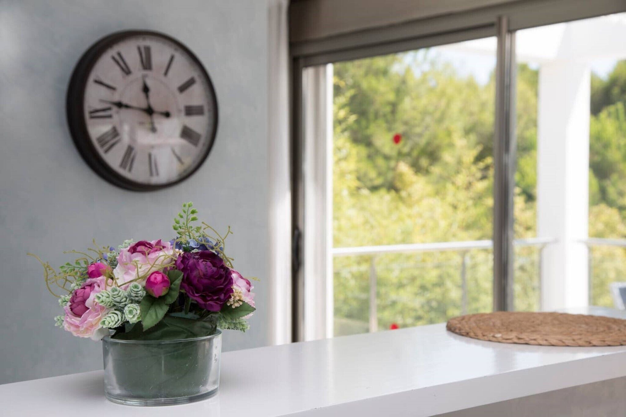 A clock on the wall, a window with a view of trees, and a vase with flowers on a white surface.
