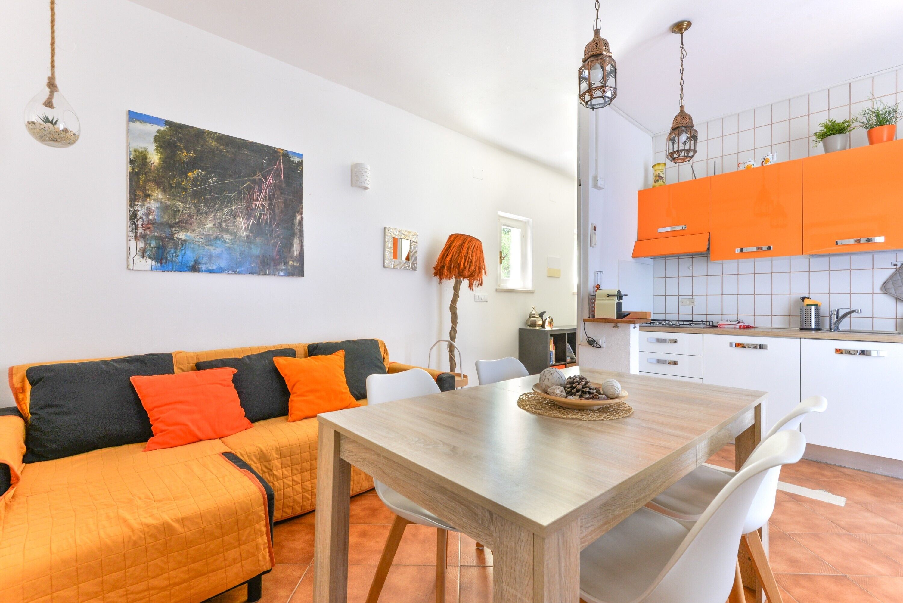 A modern living room with a wooden dining table, a sofa with orange cushions, and a kitchen with orange cabinets.