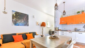 A modern living room with a wooden dining table, a sofa with orange cushions, and a kitchen with orange cabinets. - Modern apartment with garden, air conditioning and Wi-Fi (LACONA - CAPOLIVERI (LIVORNO))