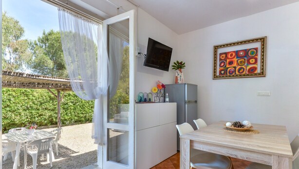 A room with a sliding glass door to a patio, a small kitchen area with a TV, a dining table with chairs, and a decorative wall piece. - Modern apartment with garden, air conditioning and Wi-Fi (LACONA - CAPOLIVERI (LIVORNO))