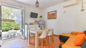 An open-plan living space with a dining area, a sofa, a TV, and a view of greenery outside. - Modern apartment with garden, air conditioning and Wi-Fi (LACONA - CAPOLIVERI (LIVORNO))