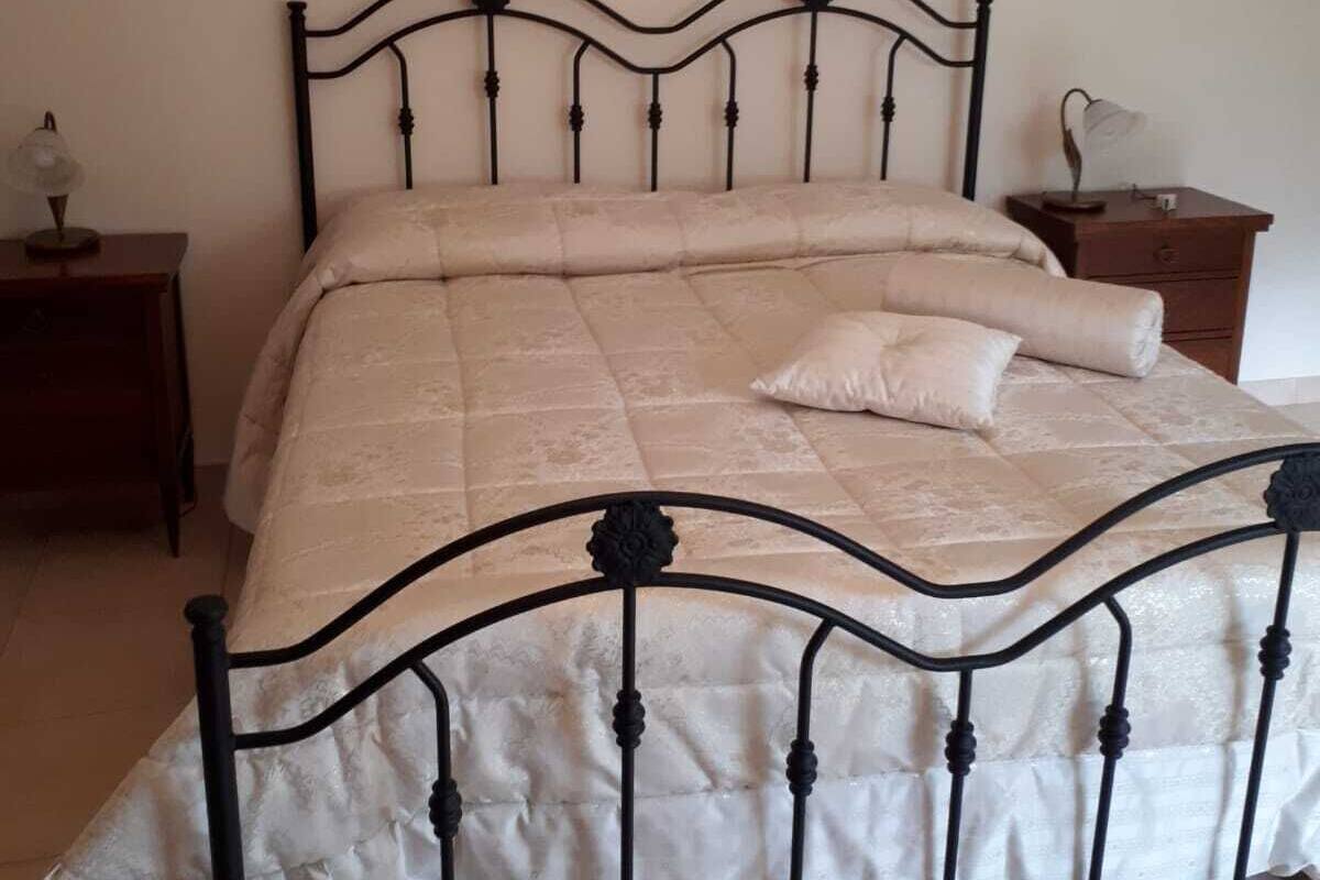 Un lit avec un cadre en métal et une couvre-lit matelassée, une table de chevet avec une lampe et un tableau encadré au mur.