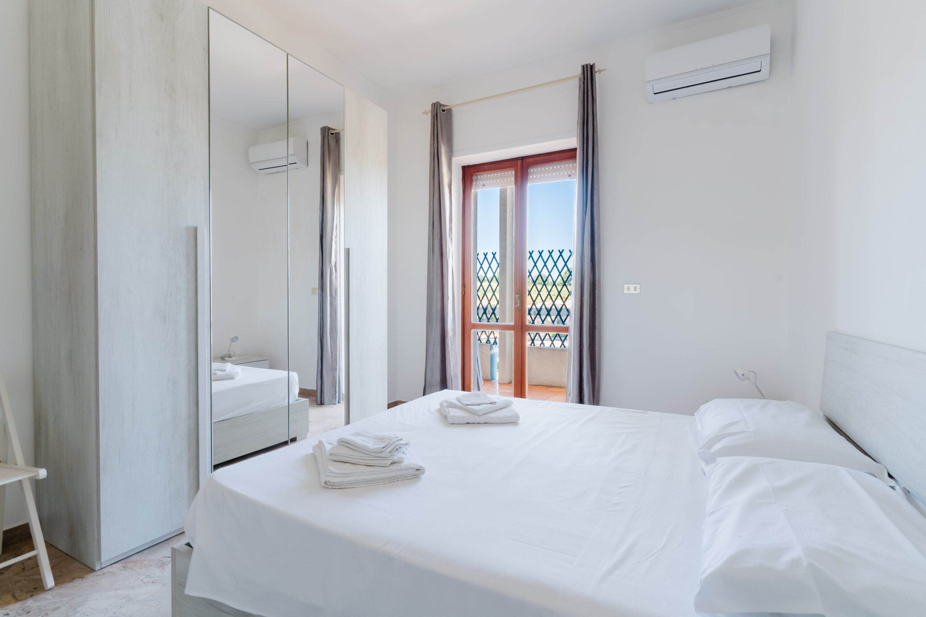 A hotel room with a bed, a mirror, an air conditioning unit, and a view of the outside.