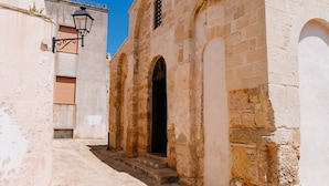 Une rue étroite bordée d’un bâtiment en pierre, avec une porte d’entrée centrale et des fenêtres en arc de cercle.
