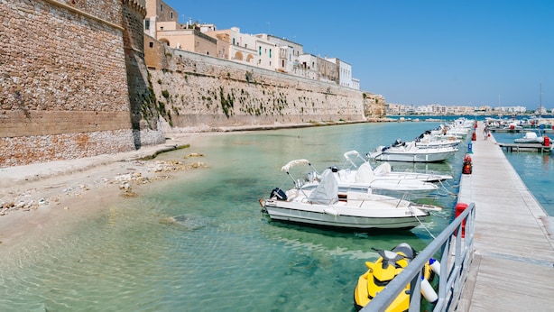 Un port de plaisance situé au bord de l’eau, avec des bateaux amarrés, un mur de pierre et des bâtiments en arrière-plan.