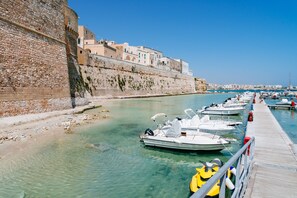 Port marítim amb embarcacions amarrades, mur de pedra i edificis al fons.