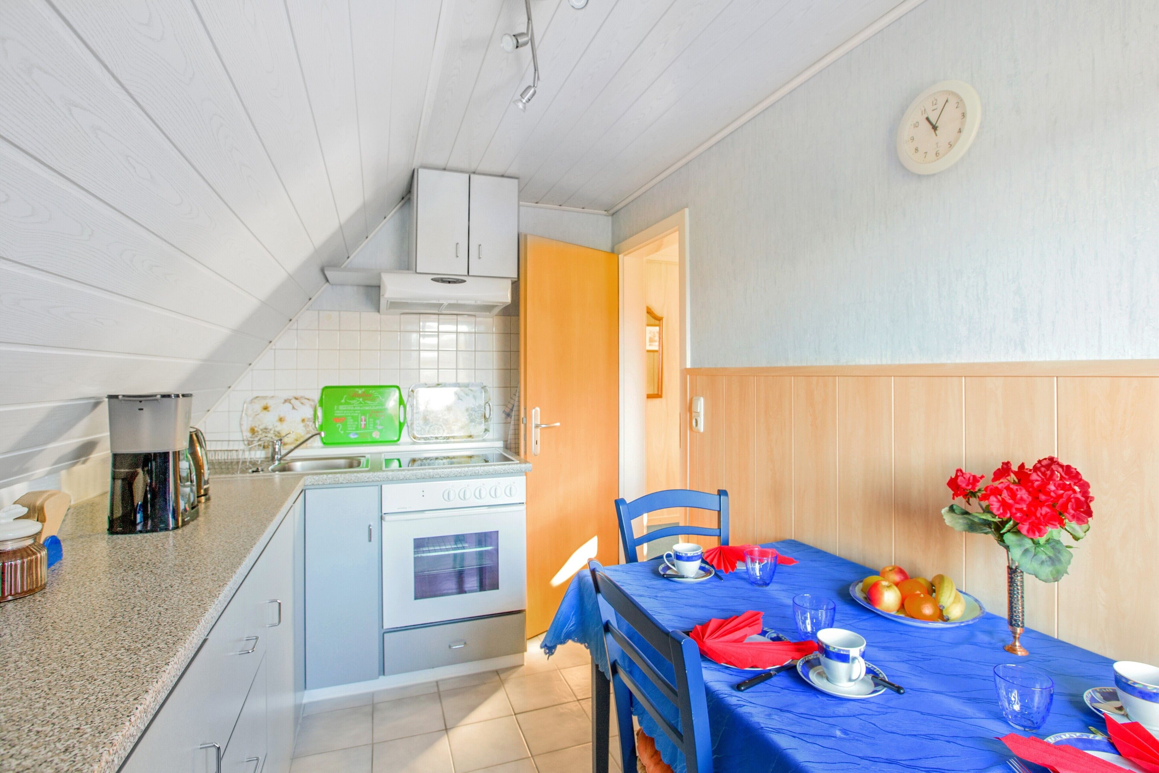 Une cuisine équipée d’une cuisinière, d’un évier et de placards, une table à manger avec des chaises, et une horloge au mur.