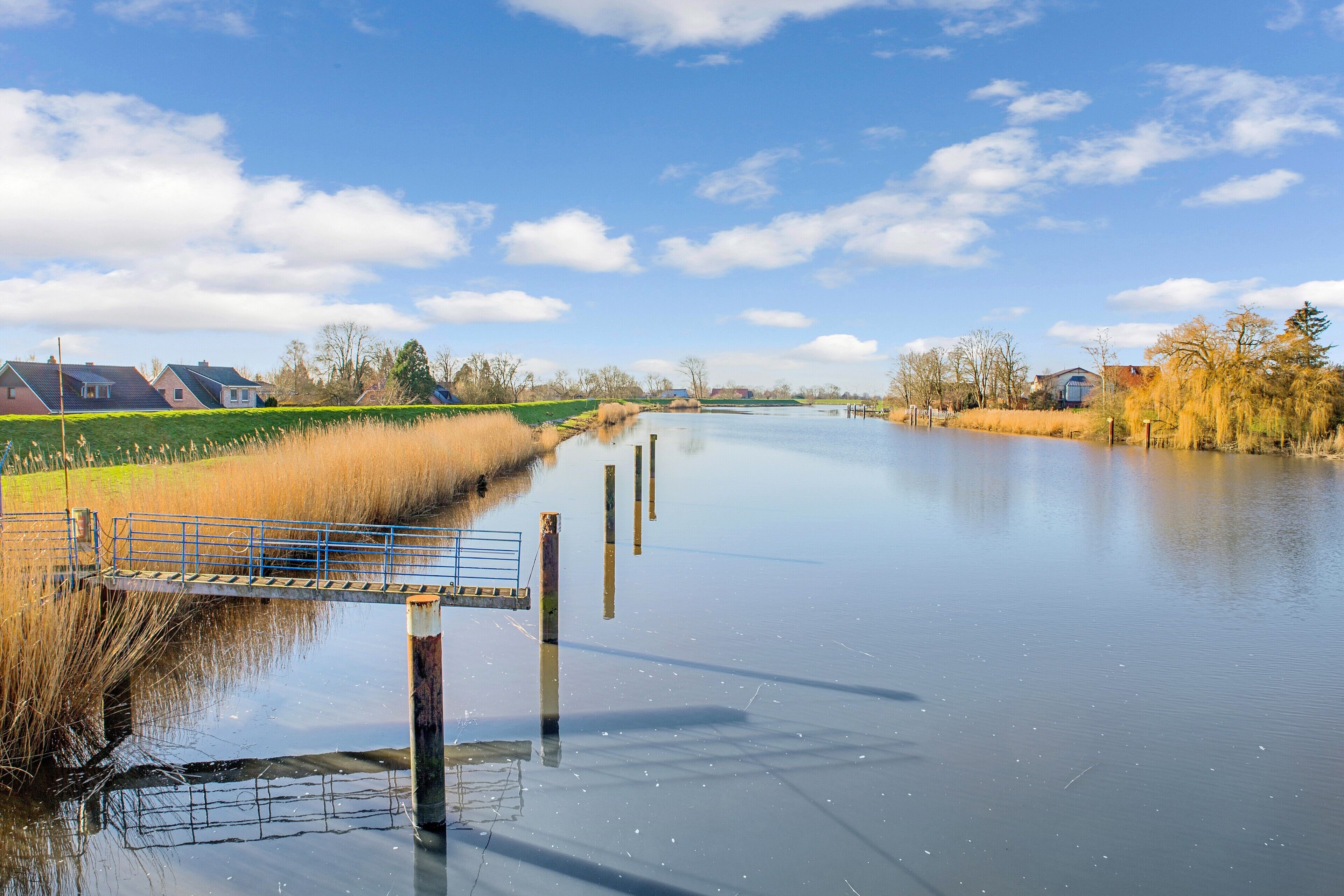 Ein ruhiger Fluss mit einer Brücke, Häusern und Bäumen im Hintergrund.
