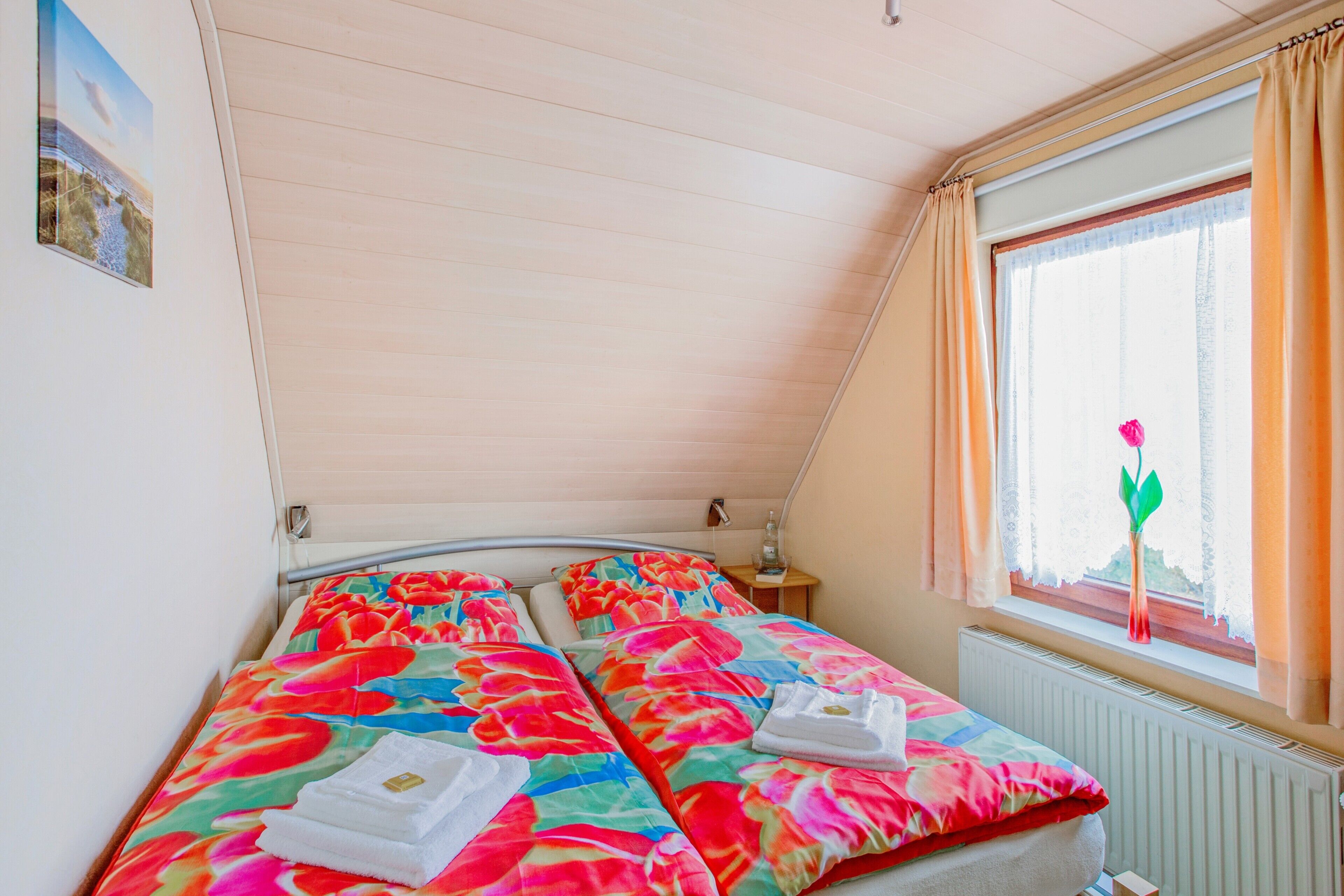 Ein Schlafzimmer mit schrägen Wänden, einem Bett mit floralem Bettzeug, einem Nachttisch mit Lampe und einem Fenster mit Vorhängen.