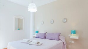 A bedroom with a bed, pillows, a hanging light fixture, a wall-mounted shelf with a lamp, and a mirror.