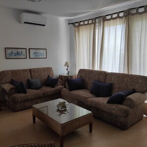 A living room with two patterned sofas, a glass coffee table, and a wall-mounted air conditioner.