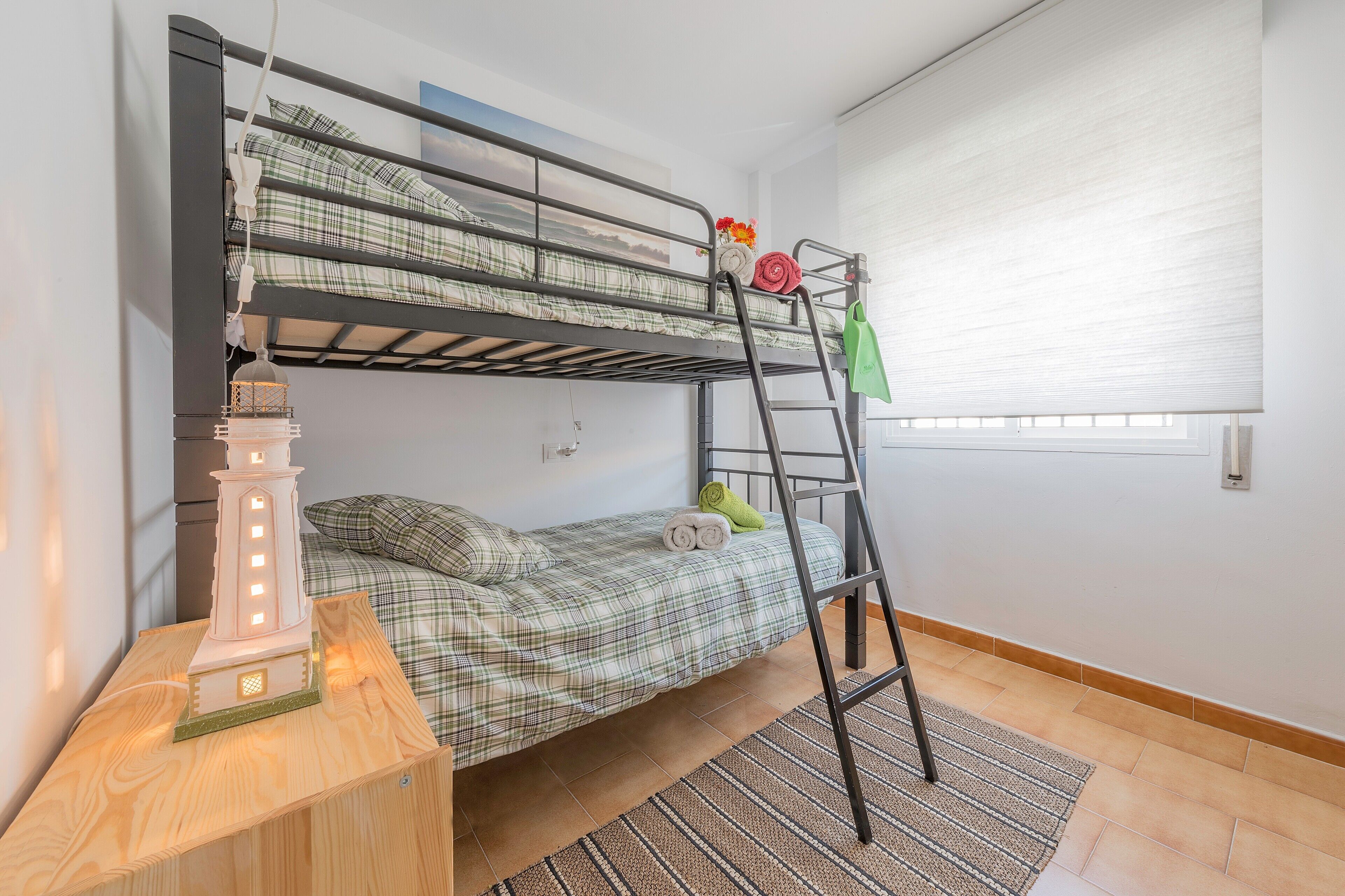 A bunk bed with a ladder, a small table with a decorative item, and a window with blinds.
