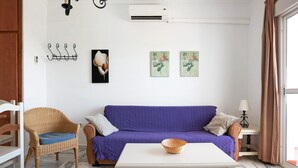 Una habitación con un sofá morado, un sillón de mimbre, una mesa blanca y un aire acondicionado instalado en la pared.