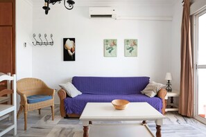 A room with a purple sofa, a wicker chair, a white table, and a wall-mounted air conditioner.