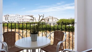 Un balcón con sillas de mimbre y una mesa, una planta en maceta y vistas de edificios al fondo.